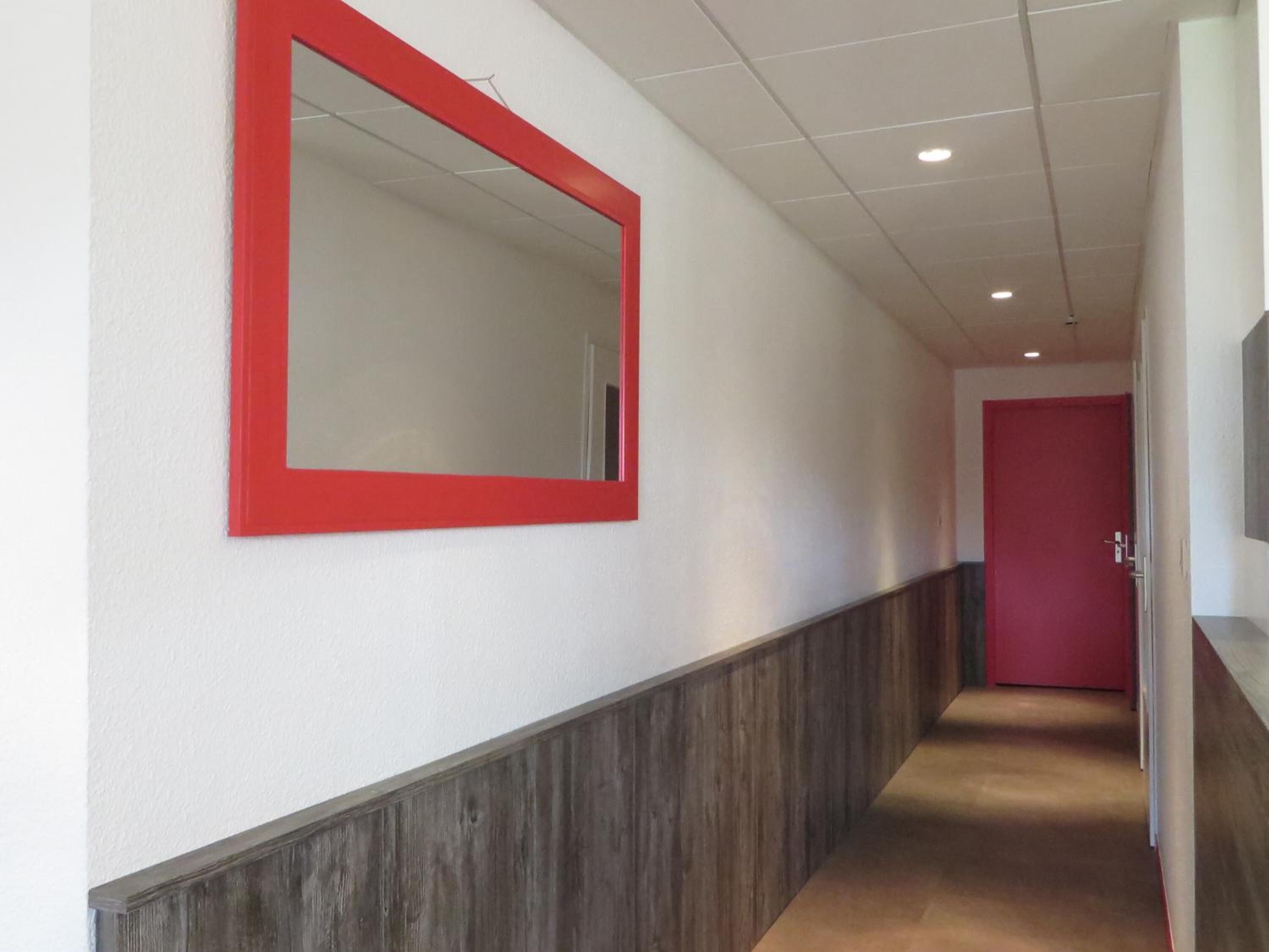 Een gang met een spiegel met rode lijst, een houten paneelwand en een rode deur aan het einde.