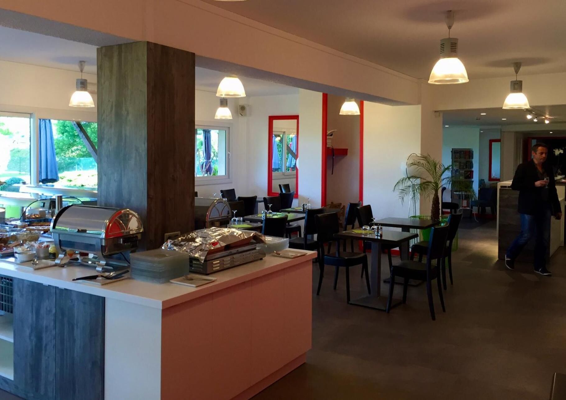 Un restaurante con mostrador central, mesas y sillas para comer. Hay una zona de buffet con comida y una parrilla. El espacio tiene un diseño moderno con iluminación colgante y ventanas amplias.