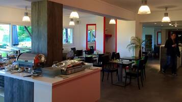 Un restaurante con mostrador central, mesas y sillas para comer. Hay una zona de buffet con comida y una parrilla. El espacio tiene un diseño moderno con iluminación colgante y ventanas amplias.