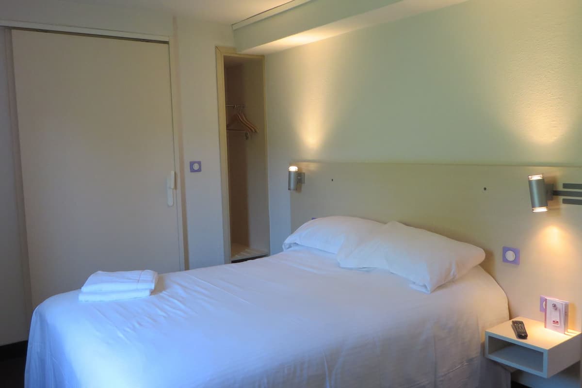 Ein Hotelzimmer mit einem Bett, einem Nachttisch mit Fernbedienung und einem Kleiderschrank mit Kleidung.