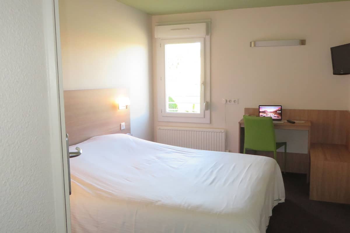 Ein Hotelzimmer mit einem Bett, einem Schreibtisch mit Computer, einem Fenster und einem an der Wand befestigten Fernseher.