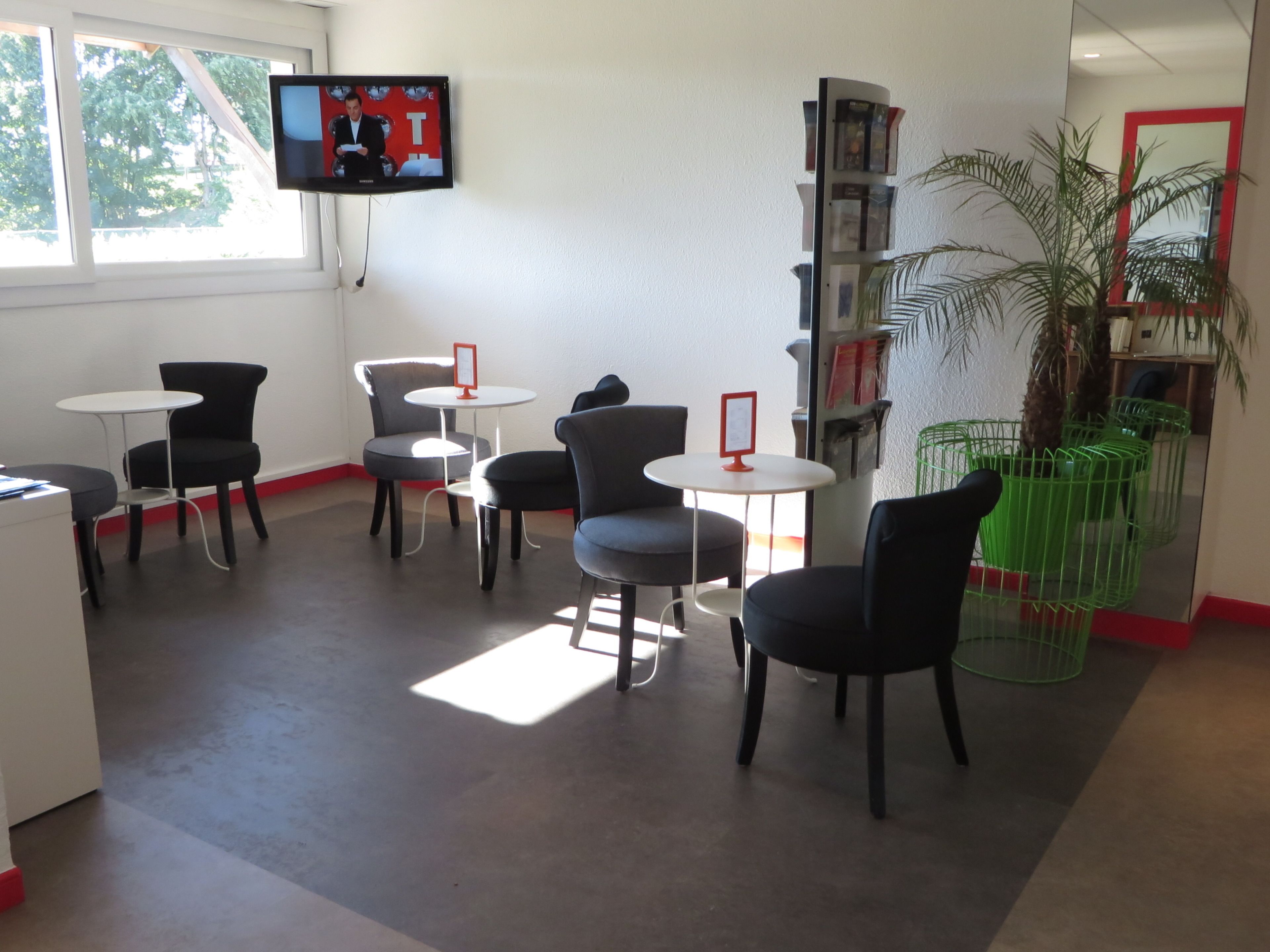 Ein modernes Zimmer mit einem wandmontierten Flachbildfernseher, mehreren schwarzen Stühlen, weißen Tischen, einem Bücherregal und einer Topfpflanze.