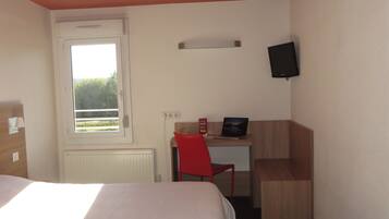A hotel room with a bed, a desk with a laptop, a red chair, a TV mounted on the wall, and a window with a view of greenery.