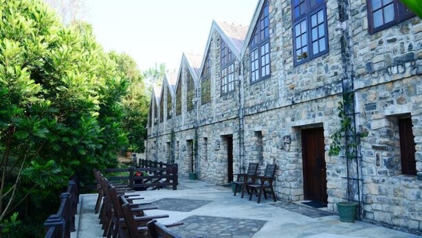Un edificio de piedra con múltiples ventanas y puertas, un patio empedrado y mobiliario de madera.