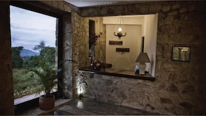 Habitación con paredes de piedra, una ventana con vistas a follaje, una lámpara colgante y una pequeña mesa con lámpara.