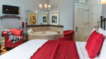 A hotel room with a bed, red and white bedding, a red armchair, a red and white patterned throw, a large mirror, a chandelier, a television, a bathtub, and a door with a white doorbell.
