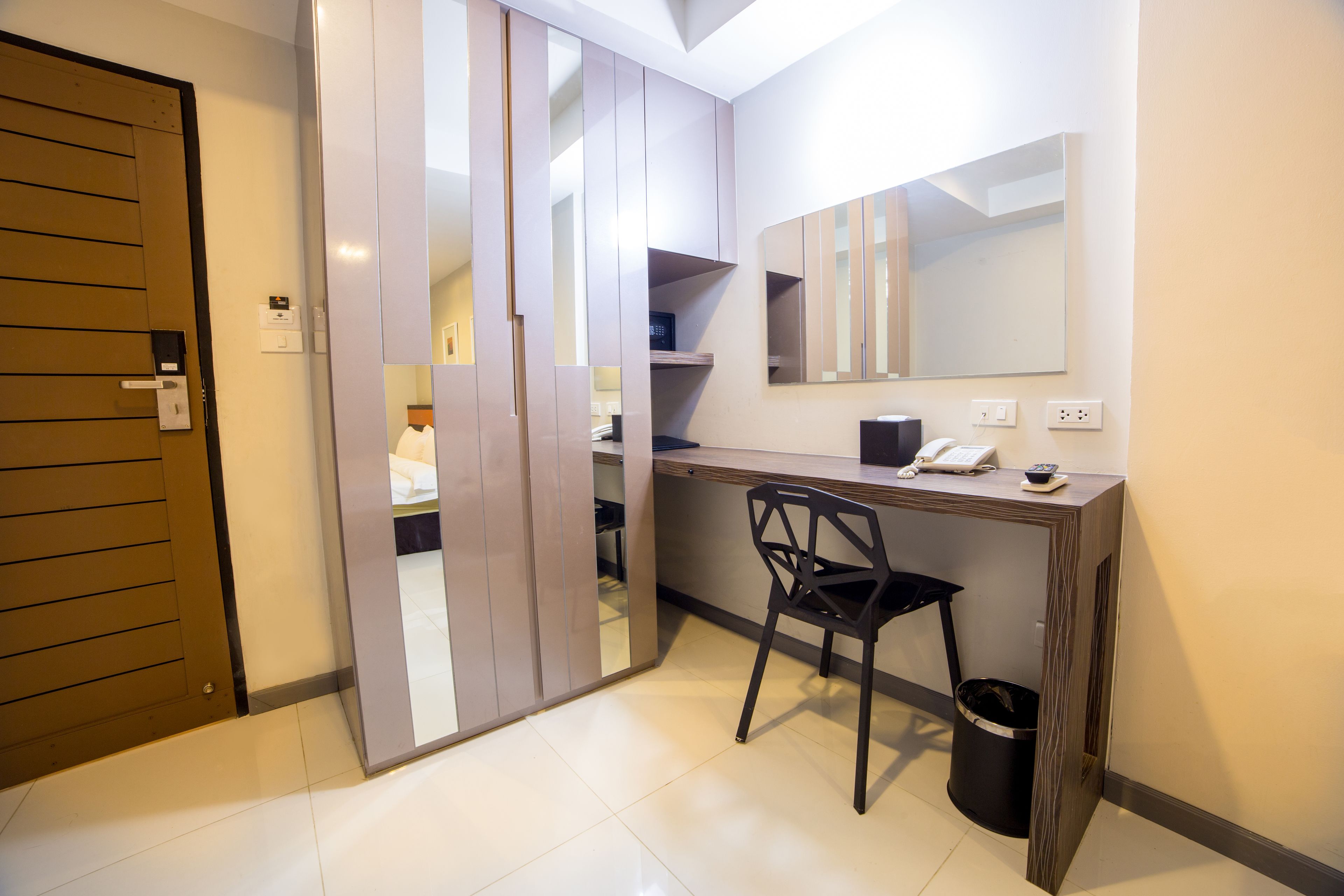 Una habitación moderna con escritorio, silla y un espejo grande. Se observa una puerta con manija y un cesto de basura.