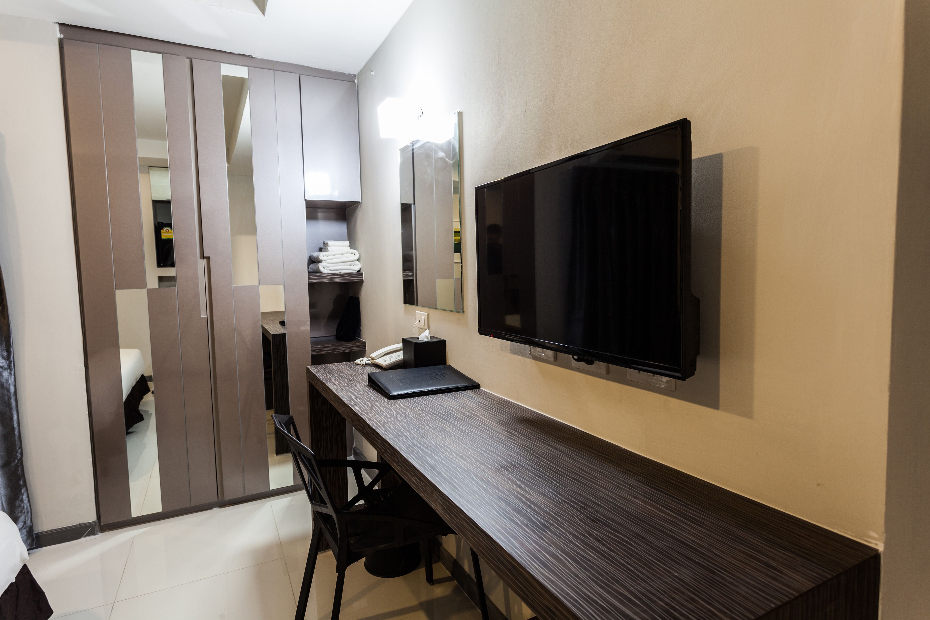 A modern hotel room with a large flat-screen TV mounted on the wall, a wooden desk with a laptop, and a wardrobe with sliding doors.