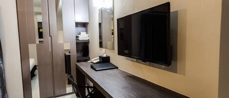 A modern hotel room with a large flat-screen TV mounted on the wall, a wooden desk with a laptop, and a wardrobe with sliding doors.
