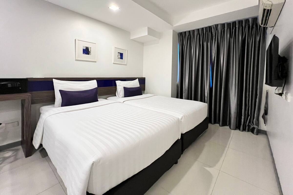 A hotel room with two beds, a flat-screen TV mounted on the wall, and a large window with dark curtains.