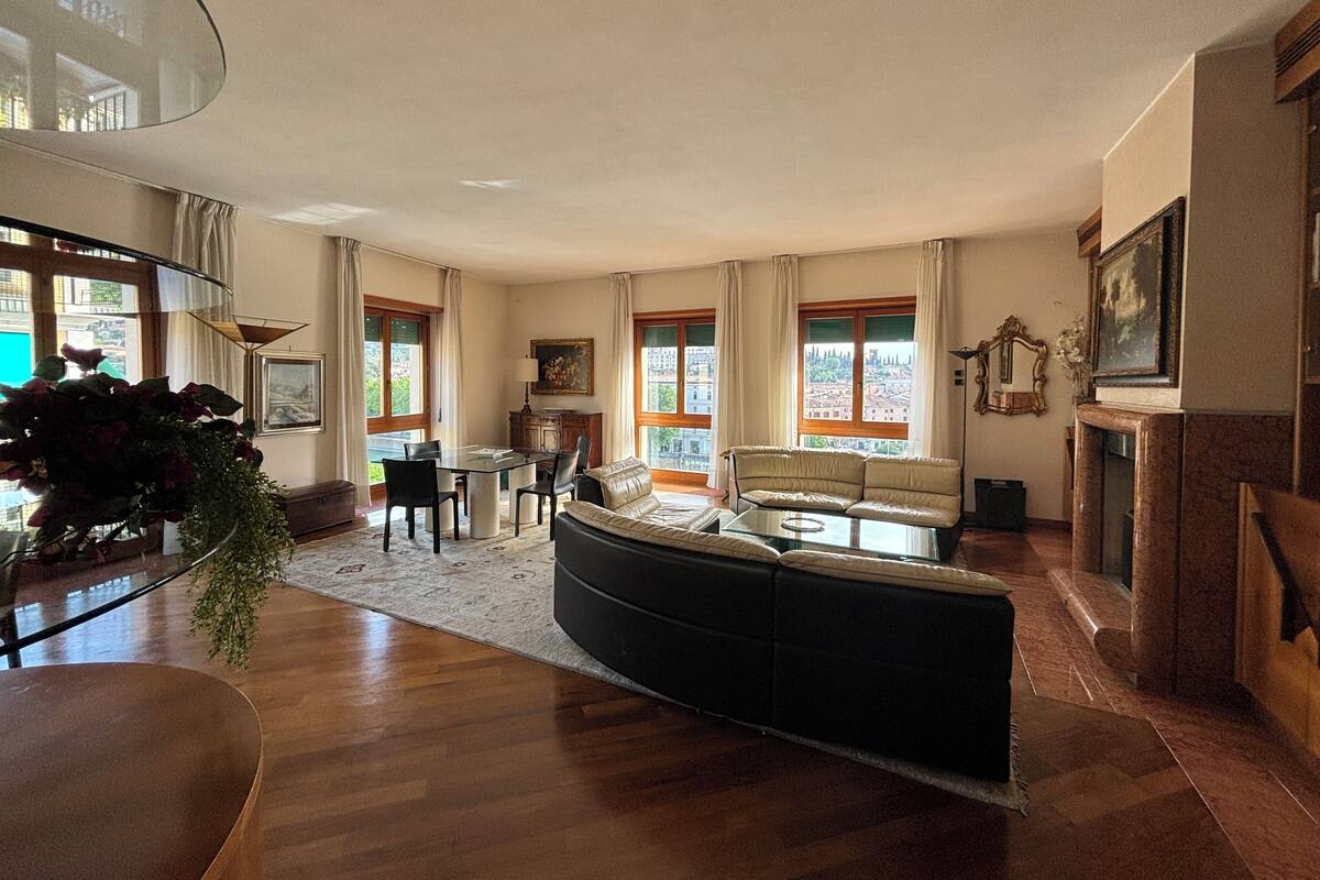 A spacious living room with a black sofa, a glass coffee table, and a dining area with a table and chairs. Large windows provide natural light and views of the city.
