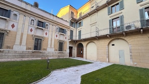 A courtyard with a paved walkway, a grassy area, and a building with classical architecture, including columns and arched doorways. - Verona Journeys (Verona)