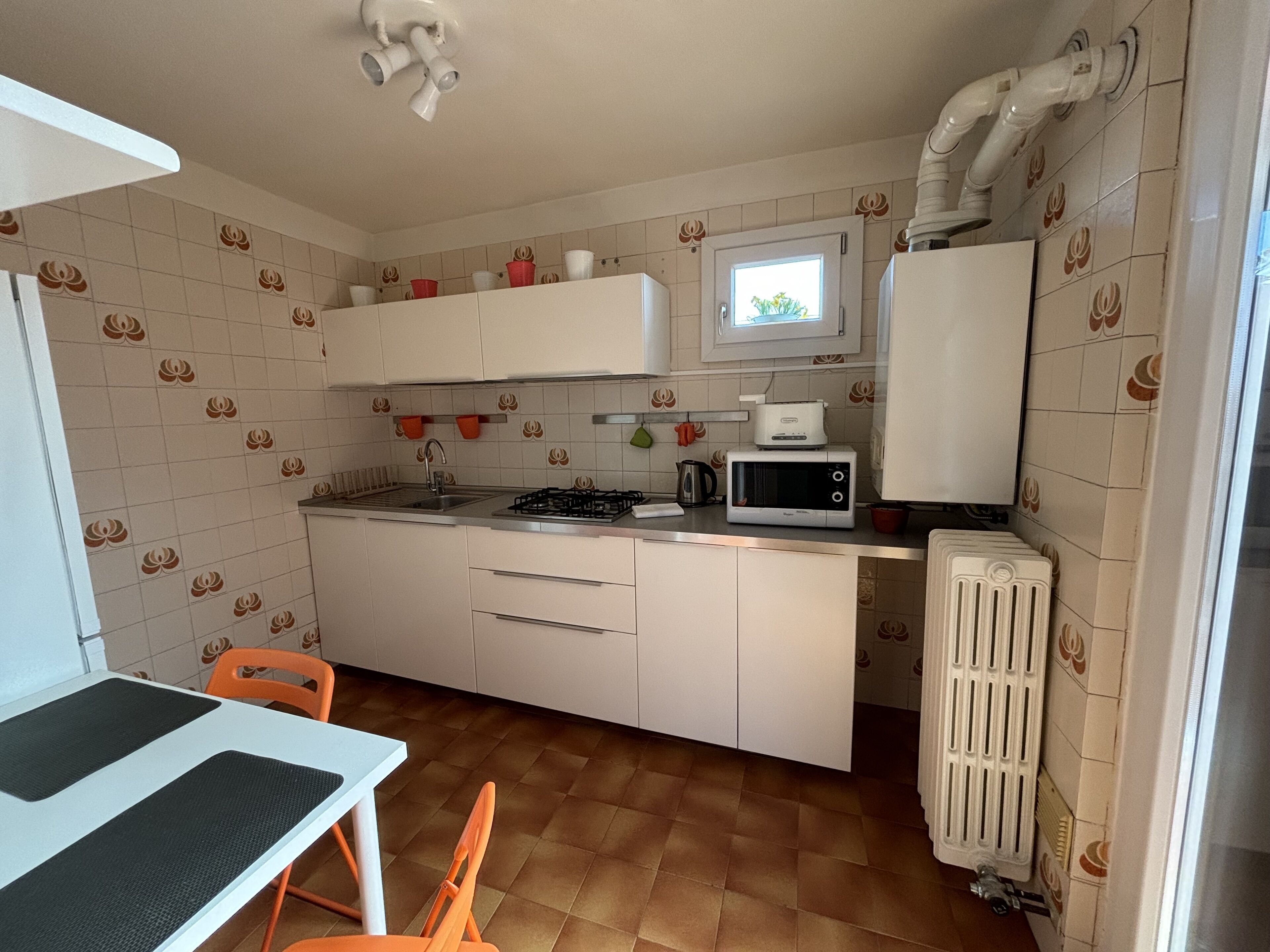 A compact kitchen with white cabinets, a microwave, a gas stove, and a sink. There is a window above the sink and a radiator next to the refrigerator.