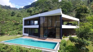 Ein modernes, zweistöckiges Haus mit Swimmingpool, umgeben von üppiger Vegetation und Hügeln.