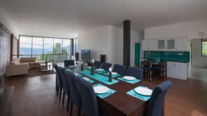 Ein moderner Essbereich mit einem langen Tisch, gedeckt für sechs Personen, umgeben von blauen Stühlen, einer Couch und einem Fernseher.
