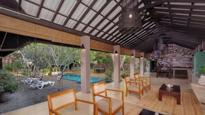 Un espacio al aire libre techado con sillas de madera, piscina y zona de comedor con un mural.