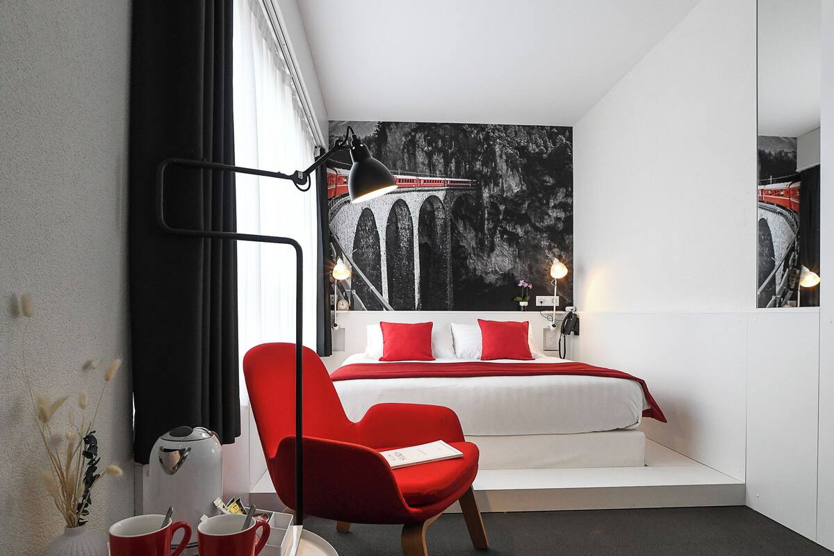 A modern hotel room with a bed, a red chair, a small table with cups, and a large window with curtains.