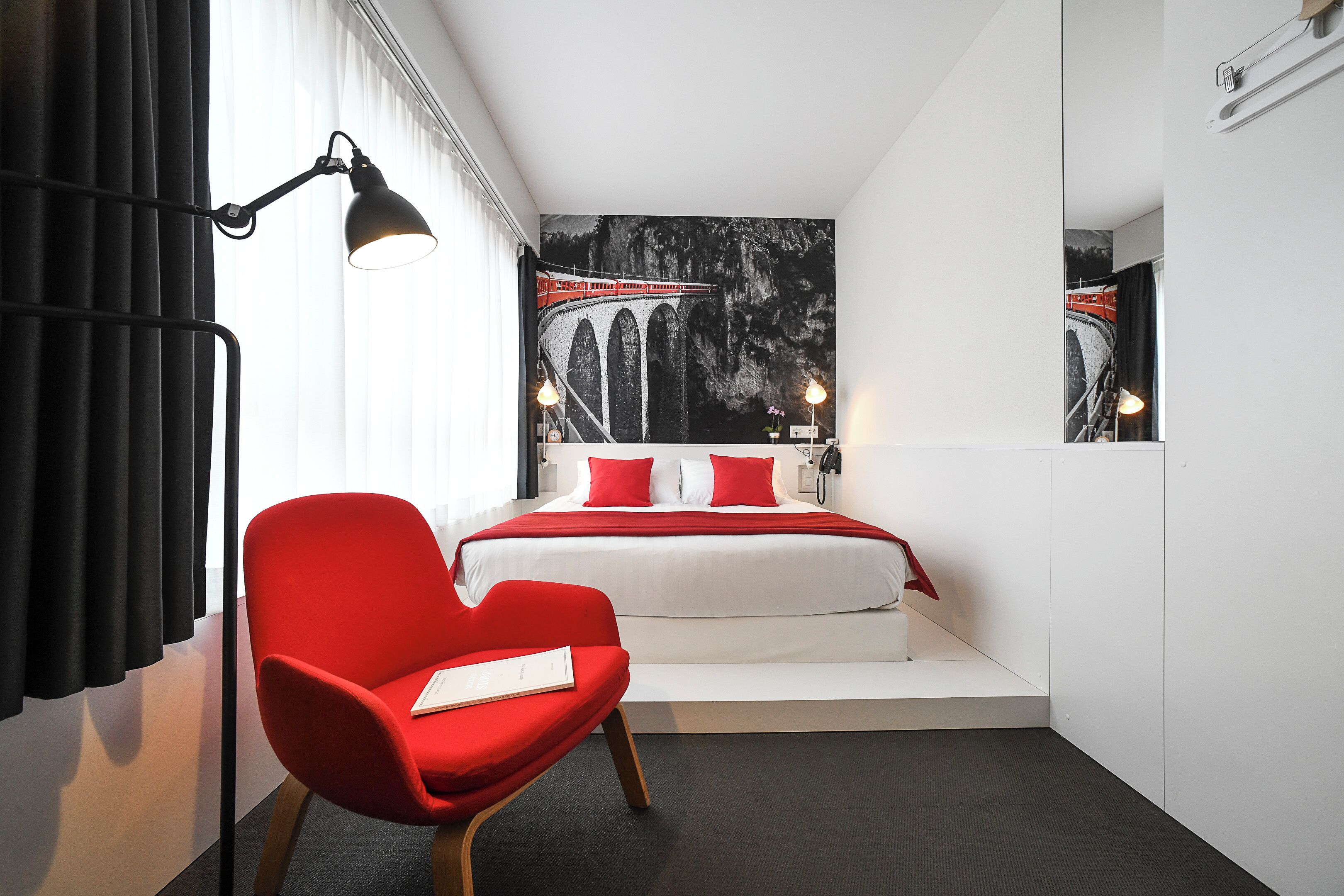 A modern hotel room with a large bed, a red chair, a black lamp, and a wall painting of a bridge.