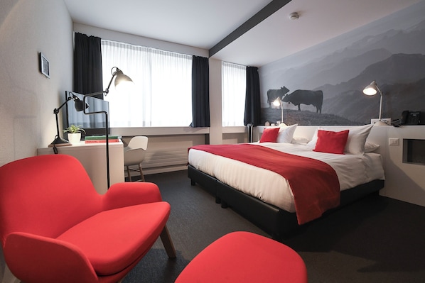 Habitación de hotel con cama, sillas rojas, un escritorio con lámpara y un mural de montañas en la pared.