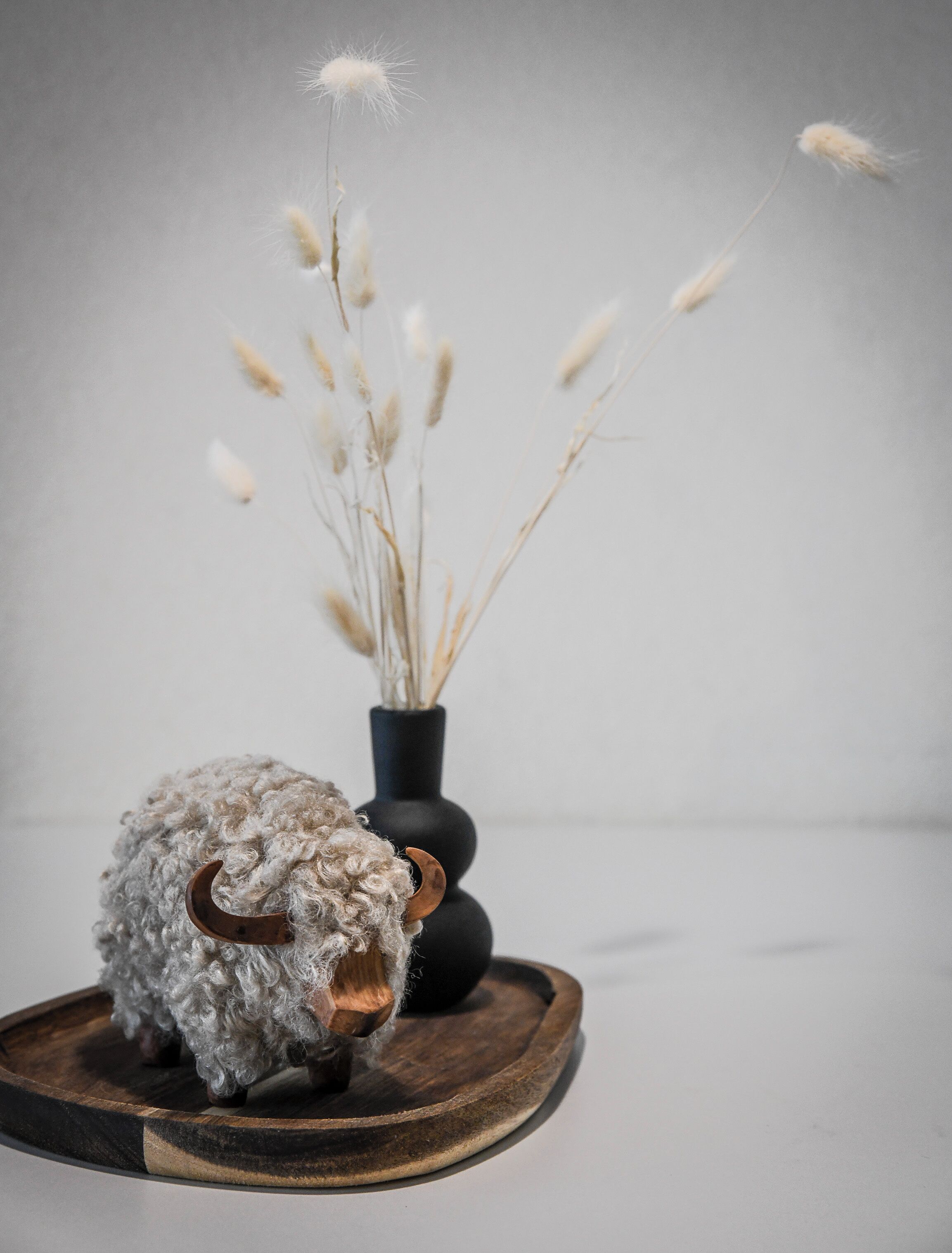 A rustic decor setup with a woolly sheep sculpture, a black vase with dried plants, and a wooden tray.
