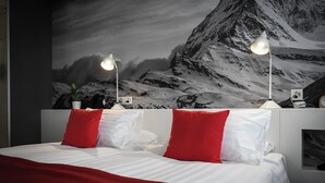 A hotel room with a bed, red and white pillows, two bedside lamps, and a mountain landscape mural on the wall. - Home Swiss Hotel (Geneva)