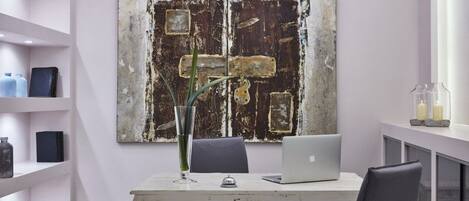 Um escritório moderno com uma obra de arte rústica na parede, uma mesa branca com um laptop e prateleiras com objetos decorativos.