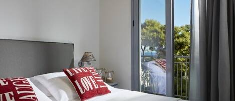 A hotel room with a large bed, a nightstand, a lamp, and a view of a balcony with trees.