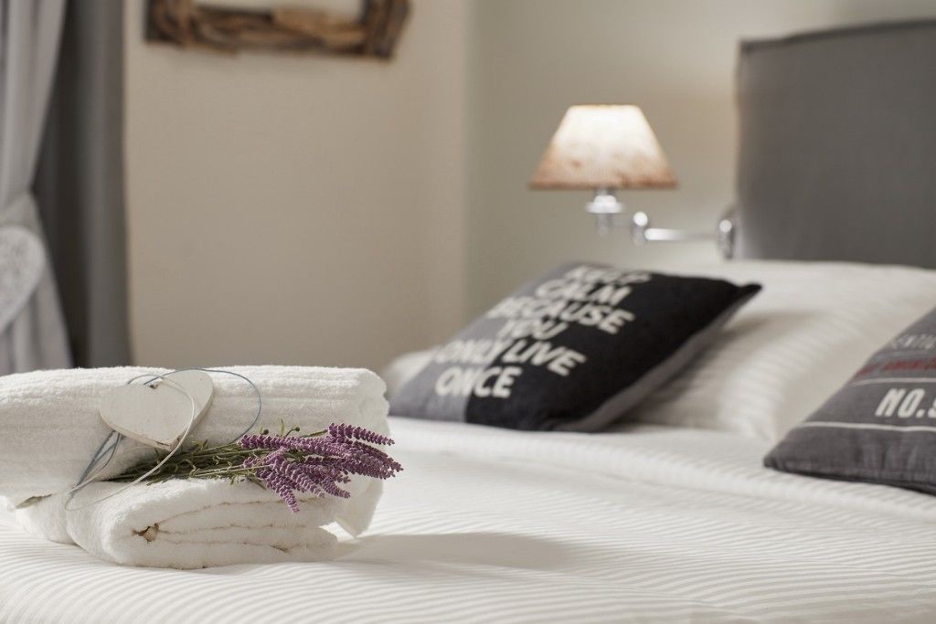 A neatly made bed with a folded white towel and a heart-shaped tag, accompanied by a sprig of lavender. There is a pillow with the text "CATHY CAN MAKE YOU LIVE ONCE" and another pillow with "NO.5" visible. A lamp is on the bedside table in the background.