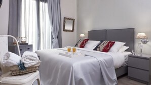 Quarto de hotel com uma cama bem arrumada, uma mesa de cabeceira com abajur, uma mesinha de apoio com um copo de água, uma cadeira com toalhas e uma janela com cortinas.