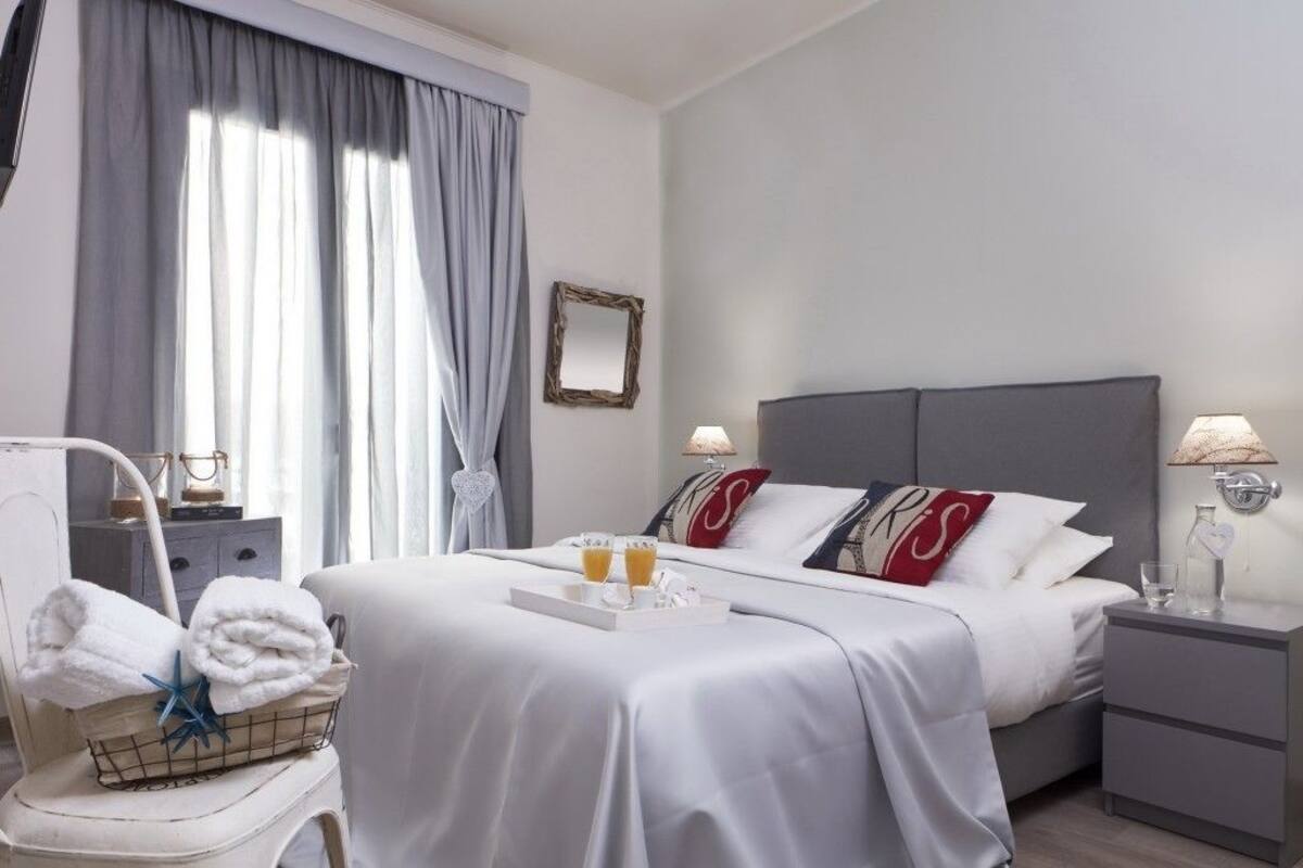 A hotel room with a neatly made bed, a bedside table with a lamp, a small bedside table with a glass of water, a chair with towels, and a window with curtains.