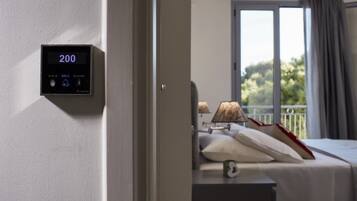 A hotel room with a digital display showing the number 200, a bed with pillows, a nightstand, and a window with a view of greenery.