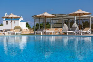 Una piscina con sillones de playa y sombrillas, un edificio blanco con una cruz, y un cielo azul despejado.