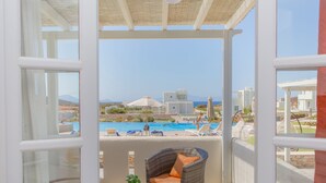 A balcony with a wicker chair and a small table, overlooking a swimming pool and buildings. - Sun and Moon Villas (Naxos)