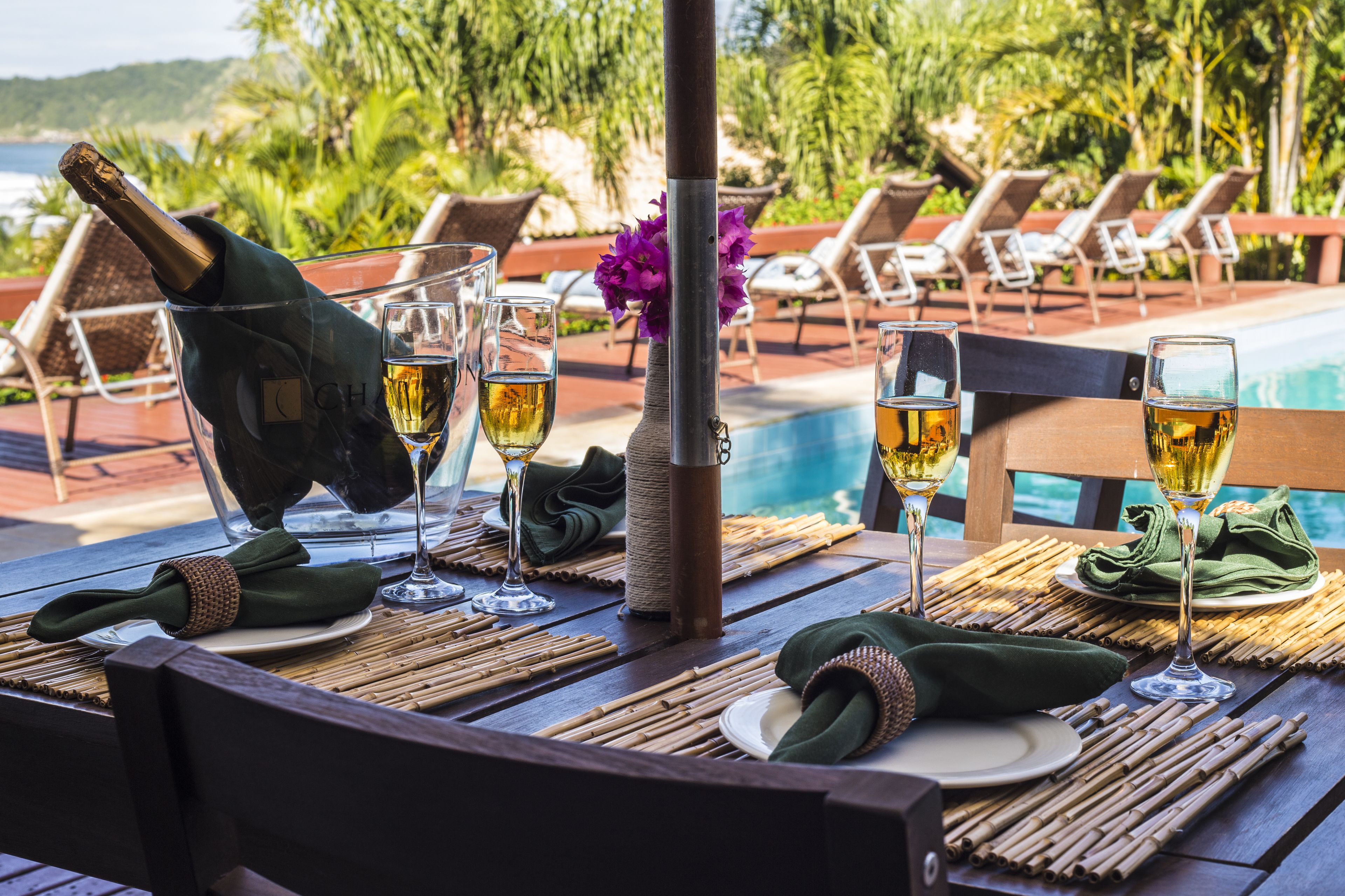 Mesa posta para refeição, com garrafa de vinho, copos e um vaso de flores, com vista para uma piscina e cadeiras de lounge.