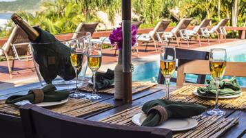 Mesa posta para refeição, com garrafa de vinho, copos e um vaso de flores, com vista para uma piscina e cadeiras de lounge.