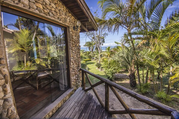 A stone building with a wooden deck and railing, overlooking a lush garden with palm trees and a view of the ocean.
