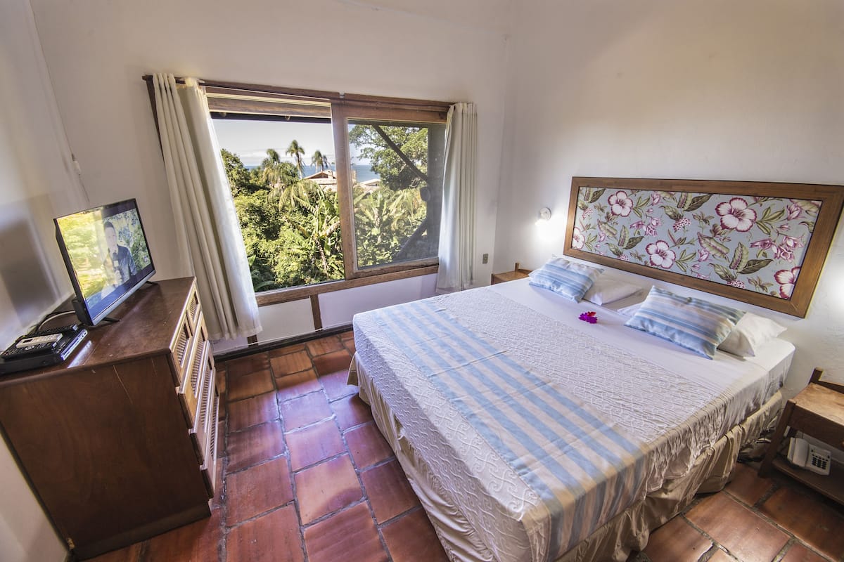 Ein Schlafzimmer mit einem großen Bett, einem Fernseher auf einem Holztisch, einem Fenster mit Vorhängen und einem blumigen Kopfstück.