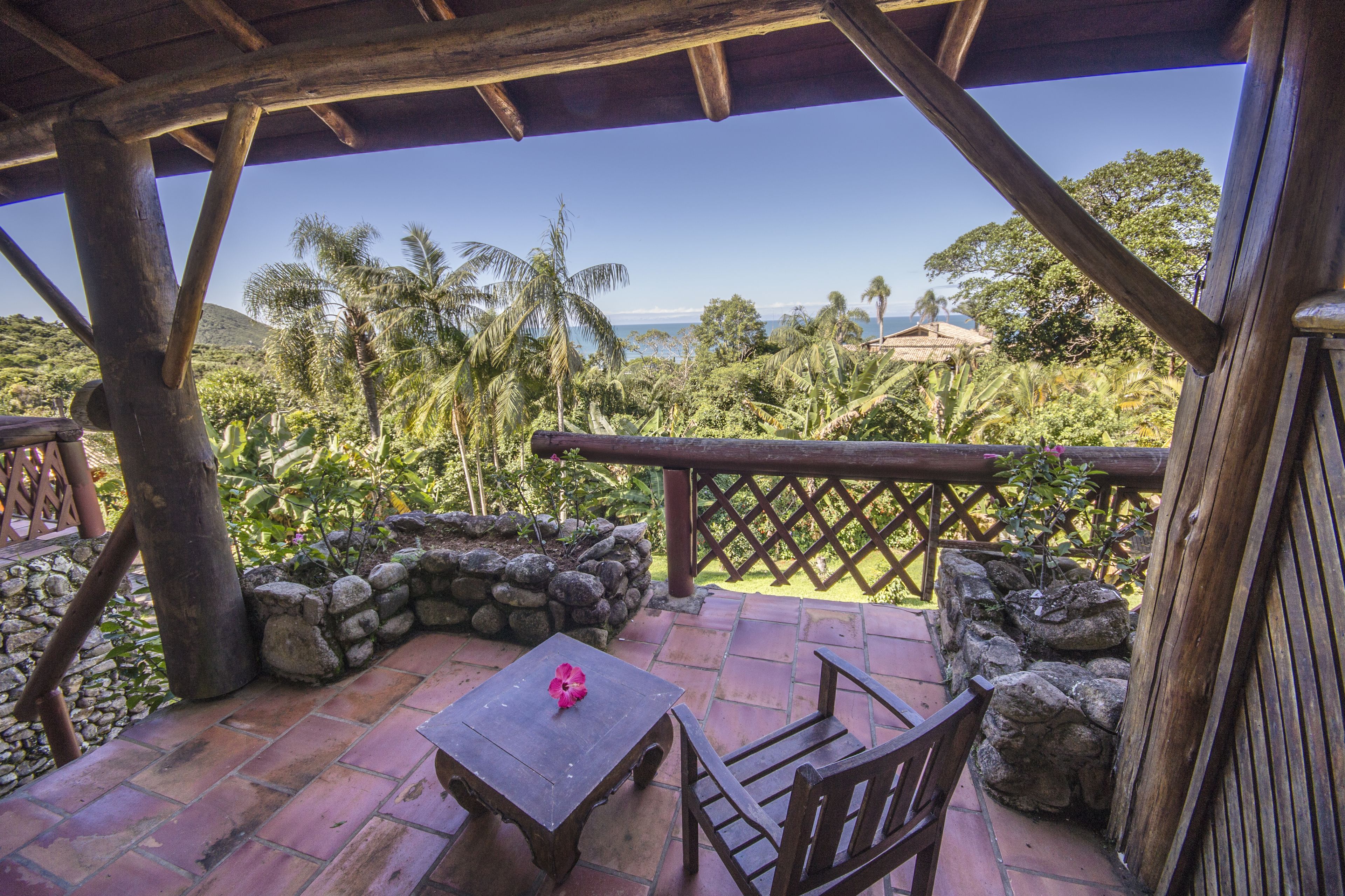 Una terraza de madera con una mesa y sillas, con vista a un paisaje tropical con palmeras y el océano.