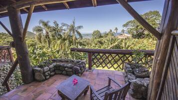 Una terraza de madera con una mesa y sillas, con vista a un paisaje tropical con palmeras y el océano.
