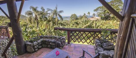 Una terraza de madera con una mesa y sillas, con vista a un paisaje tropical con palmeras y el océano.
