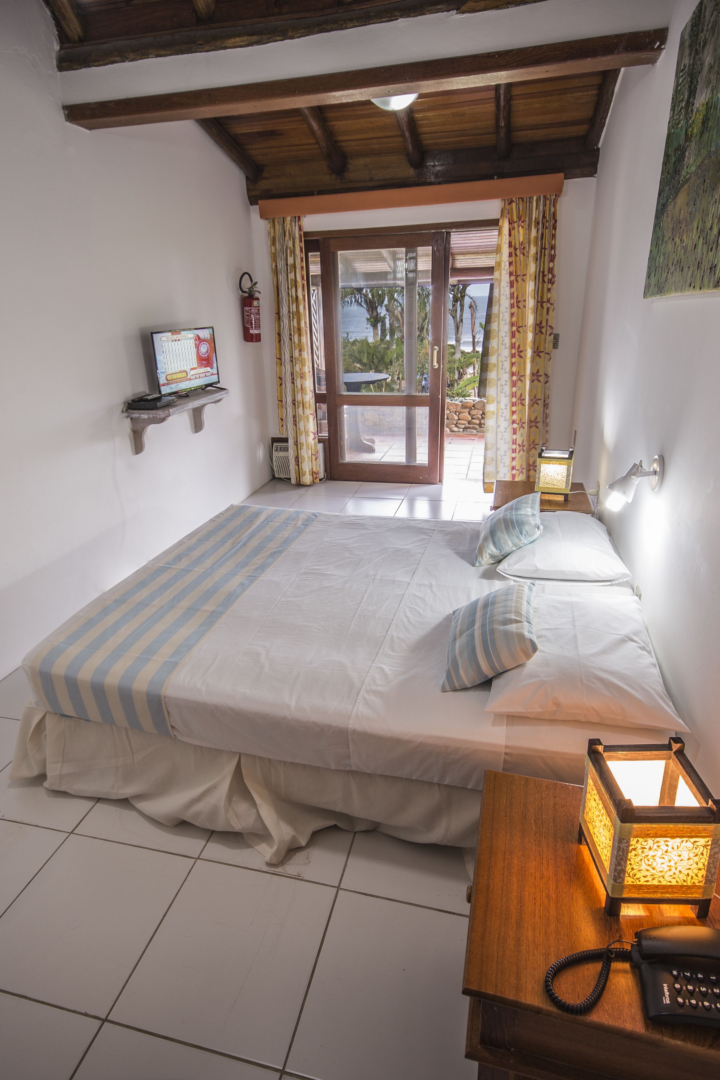 Un dormitorio con cama, una mesita de noche con lámpara, un extintor y una ventana con cortinas.