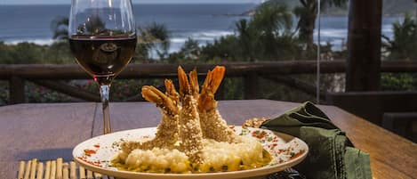 A plate of food with shrimp on a wooden table, a glass of red wine, and a view of the ocean.