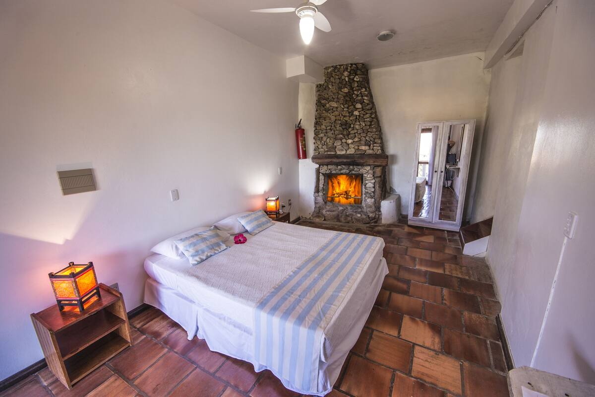 Ein Schlafzimmer mit Kamin aus Stein, einem Bett mit gestreifter Bettdecke und einem Nachttisch mit Laterne.