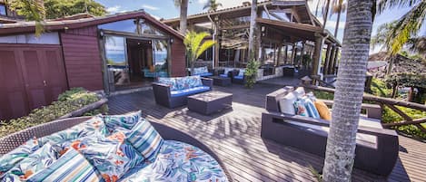 Una terraza de madera con mobiliario exterior, palmeras y una zona cubierta con vista al océano.