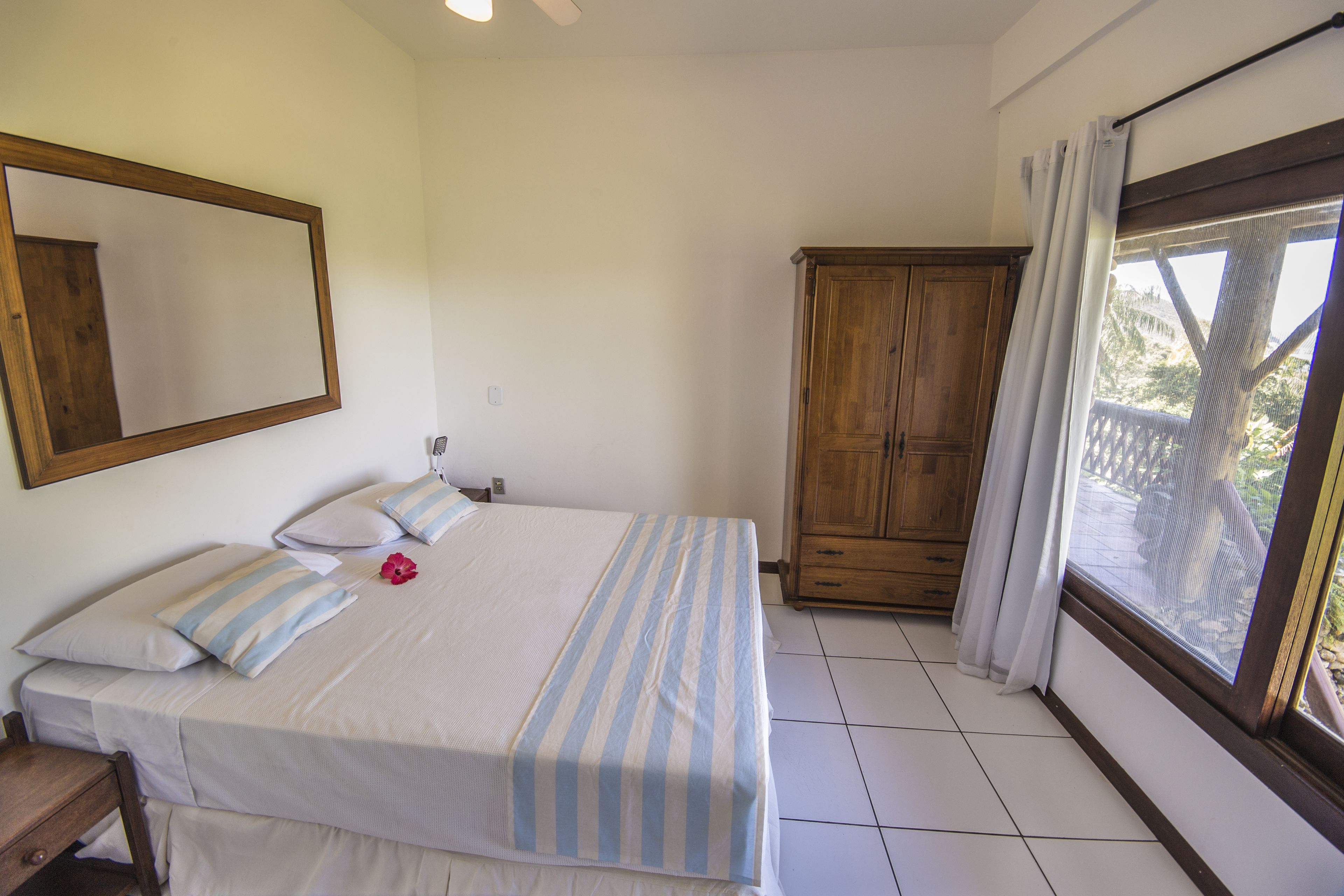 Een slaapkamer met een bed, een houten kast, een raam met gordijnen en een spiegel aan de muur.
