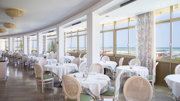 Ein Restaurant mit gedeckten Tischen, großen Fenstern mit Blick auf den Strand und Korbstühlen mit grünen Kissen.