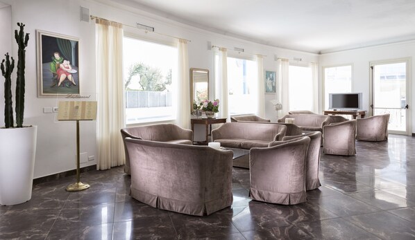A spacious living area with multiple seating options, a flat-screen TV, and a large window providing natural light. - Hotel Acapulco (Cervia)