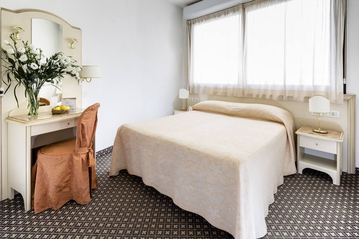A hotel room with a bed, a desk with a chair, a mirror, a lamp, and a window with curtains.