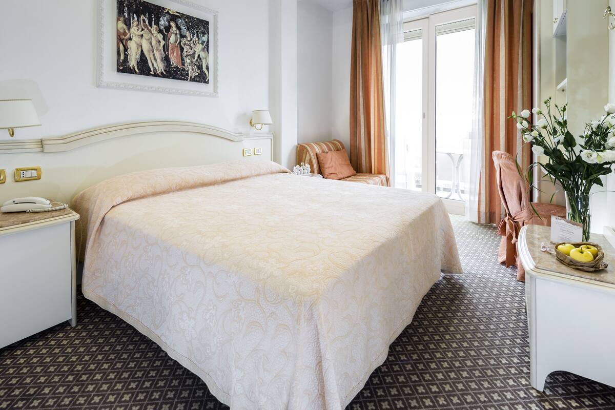 A hotel room with a large bed, a desk with a telephone, a chair, a vase with flowers, and a painting on the wall.
