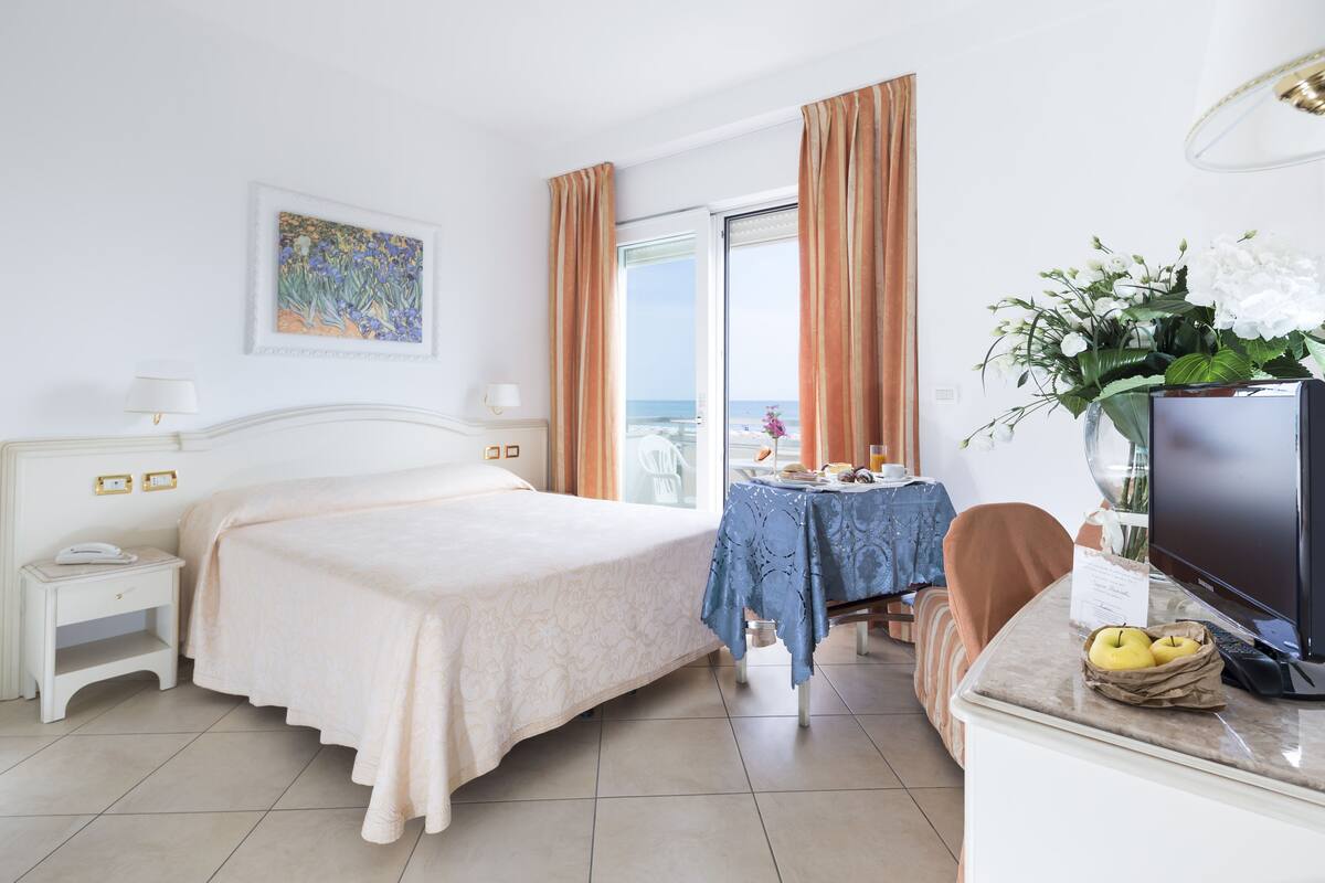 A hotel room with a bed, a desk with a computer, a chair, a small table with a vase of flowers, and a view of the sea through a window.